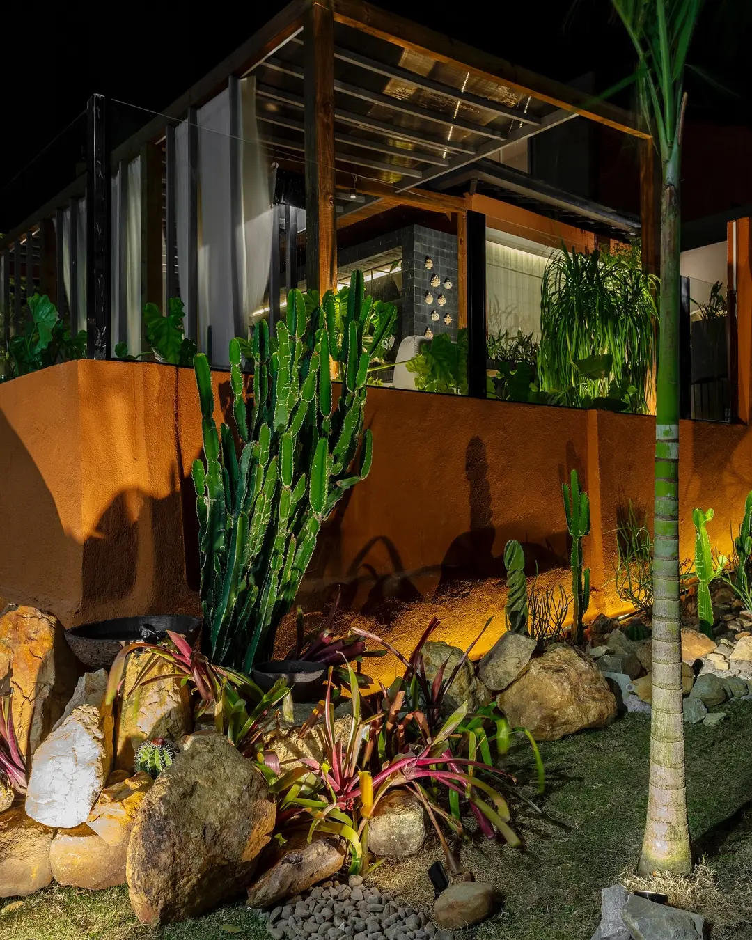 Vista noturna do ambiente Passeio para a Casa, com jardim de cactos, pedras naturais e iluminação cênica.